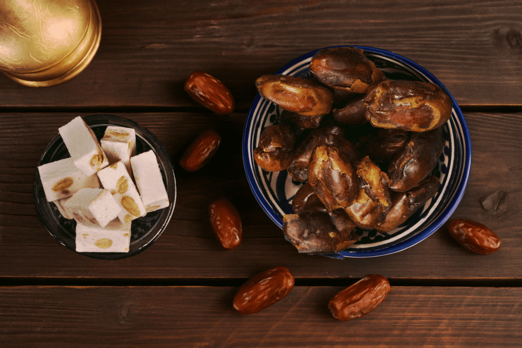 A ceramic bowl filled with glossy pitted dates on a rustic wooden table, surrounded by loose dates and traditional sweets.